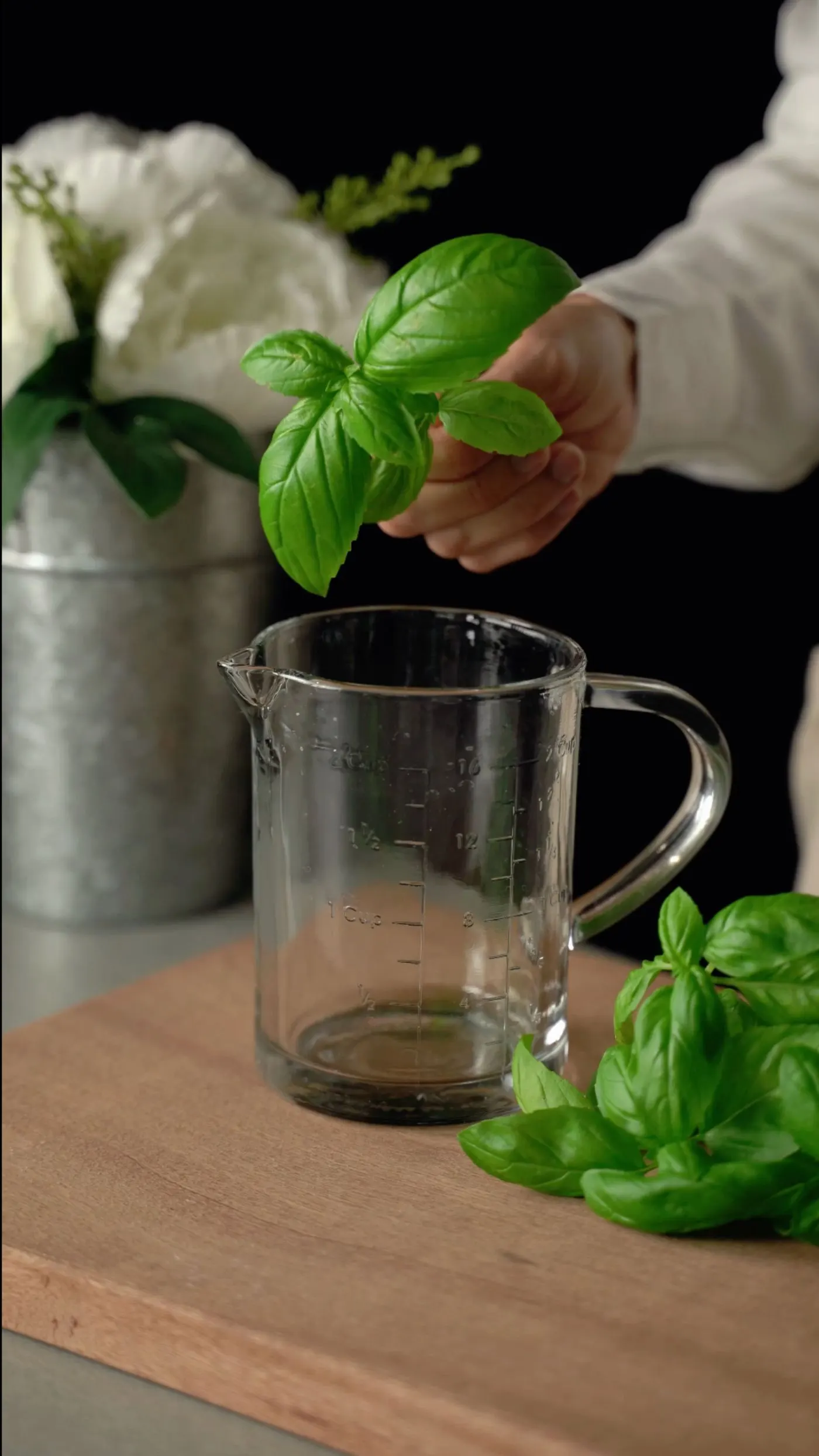 A hand dropping fresh basil leaves into a glass measuring jug to make an herbal syrup